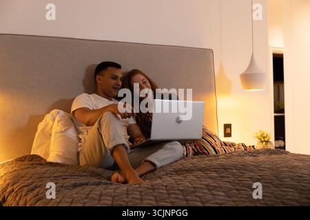 Giovane coppia che si rilassa sul letto, guarda il portatile e goditi una serata accogliente insieme Foto Stock