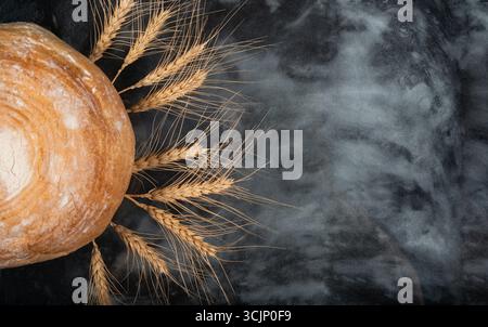 Il pane rotondo appena sfornato riposa accanto a punte di grano dorato su una superficie scura, mostrando un'estetica invitante e rustica da forno. Foto Stock