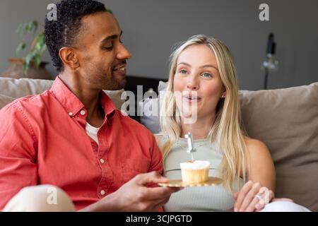 Coppia che festeggia a casa, gustando un cupcake con espressioni gioiose sul divano Foto Stock