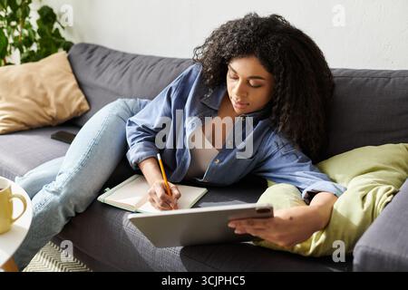 Una giovane donna lavora come freelance mentre si rilassa sul suo divano in un appartamento moderno. Foto Stock