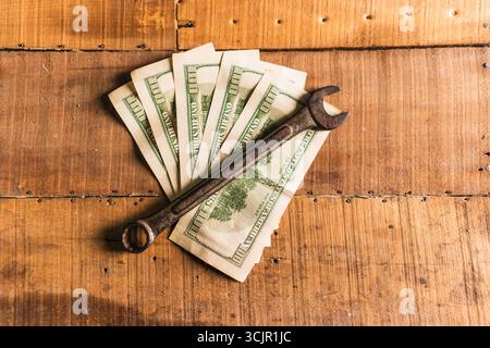 Guanti da lavoro in pelle con una chiave arrugginita e dollari americani su un tavolo da lavoro rustico in legno Foto Stock