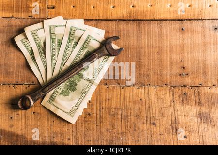 Guanti da lavoro in pelle con una chiave arrugginita e dollari americani su un tavolo da lavoro rustico in legno Foto Stock