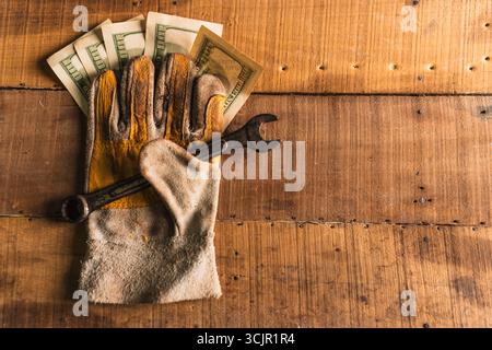 Guanti da lavoro in pelle con una chiave arrugginita e dollari americani su un tavolo da lavoro rustico in legno Foto Stock