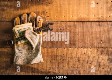 Guanti da lavoro in pelle con una chiave arrugginita e dollari americani su un tavolo da lavoro rustico in legno Foto Stock