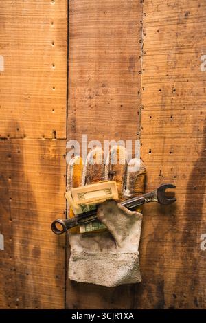 Guanti da lavoro in pelle con una chiave arrugginita e dollari americani su un tavolo da lavoro rustico in legno Foto Stock