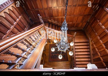 Guardando in basso la grande scala in legno e il lampadario all'interno di Knightshayes Court, Tiverton, Devon, Regno Unito, il 7 settembre 2025 Foto Stock