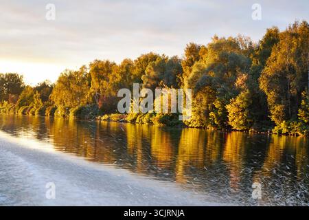 Tramonto dorato sul fiume con spettacolari nuvole e riflessi luminosi sull'acqua. Gli alberi autunnali illuminati dalla calda luce serale creano una natura tranquilla Foto Stock