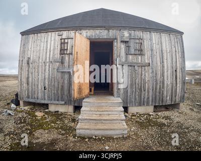 La stazione di Kinnvika Nordaustlandet Svalbard // NORDEAUSTLANDET, Svalbard - la stazione di Kinnvika, un avamposto storico di ricerca, è raffigurata a Nordaustlandet nelle Svalbard e Jan Mayen. La stazione, costruita con tavole di legno intemperie, presenta un'entrata prominente con una porta di legno aperta e una serie di scale che vi conducono. Piccole finestre con persiane sono visibili sulla facciata curva dell'edificio. Il paesaggio circostante è arido e roccioso, caratteristico dell'ambiente artico. La stazione di Kinnvika servì come base per spedizioni scientifiche in questa remota regione. Foto Stock