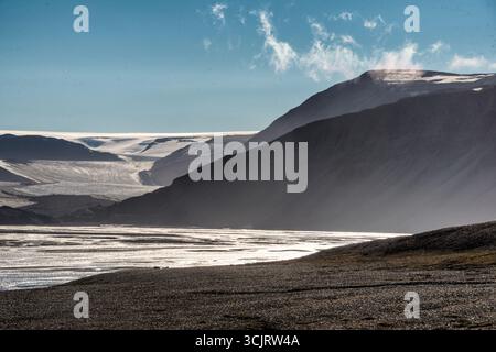 Paesaggio glaciale di Faksevagen Spitsbergen Svalbard | SPITSBERGEN, Svalbard — Una lingua glaciale scende tra creste montuose scure verso la pianura di acqua di fusione di Faksevagen, la sua superficie luccica sotto un cielo polare limpido. Spitsbergen, la più grande isola dell'arcipelago di Svalbard, detiene circa il sessanta per cento della sua superficie sotto il ghiaccio, e ghiacciai come questo alimentano i ruscelli di acqua di fusione intrecciati visibili in primo piano. I tassi di ritiro qui sono tra i più rapidi mai registrati nell'Artico. Foto Stock