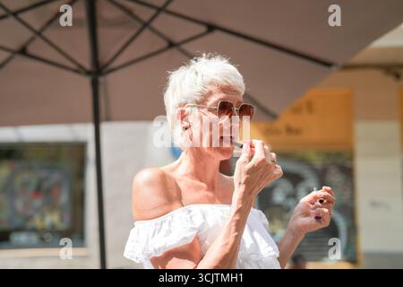Una donna con una camicia bianca sta indossando il rossetto. Lei è sotto un ombrello. L'ombrello è grande e offre ombra dal sole Foto Stock