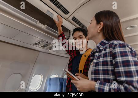 Una giovane coppia si prepara per un volo su un aereo. L'uomo mette i bagagli nel cestino sospeso mentre la donna sorride e tiene i passaporti, entrambi appea Foto Stock