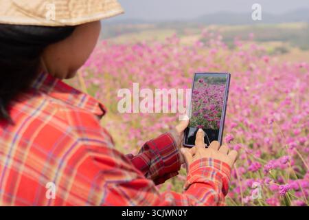 Vista posteriore di una donna turistica corta e irriconoscibile che scatta foto di montagna e campo di fiori e cielo limpido con uno smartphone. Foto Stock