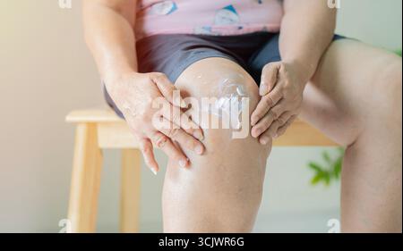 Donna anziana che applica crema al ginocchio dolore in salotto, Self-Treatment, Healthcare at Home Concept. Foto Stock