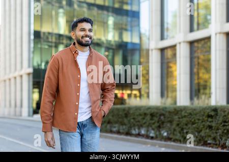 Giovane uomo d'affari indiano con la barba che cammina tranquillamente vicino ai moderni edifici di uffici, sorridendo mentre guarda lontano, godendo l'atmosfera vibrante di una carriera di successo e delle aspirazioni future Foto Stock