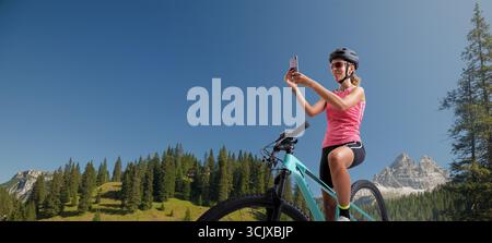Una giovane allegra donna in abbigliamento sportivo siede sulla sua mountain bike in un paesaggio di montagna, sorridendo mentre scatta una foto del paesaggio mozzafiato con Foto Stock