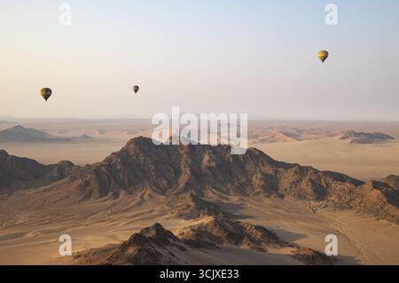Tre mongolfiere sopra una pianura arida e isolate creste di montagna ai margini del deserto del Namib. Vista aerea da un'altra mongolfiera. Namibia Foto Stock