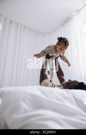 Felice papà africano che solleva la sua simpatica figlia birazziale mentre si sdraia sul letto in una luminosa camera bianca. Concetto di famiglia interrazziale. Foto Stock