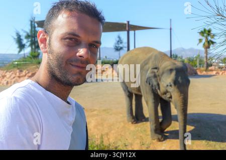 Uomo sorridente con elefante sullo sfondo allo zoo Attica di Atene, Grecia. Foto Stock