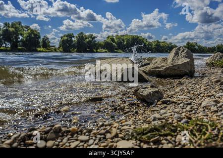 L'acqua spruzza sulle pietre sulle rive del Danubio in una giornata limpida vicino a Bogen, nella bassa Baviera. Il sole splende e le nuvole sono riflesse Foto Stock