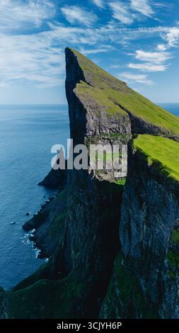 Le maestose scogliere si innalzano nettamente dalle tranquille acque del Nord Atlantico nelle Isole Faroe. Lussureggianti colline verdi contrastano con gli affioramenti rocciosi, creando un panorama naturale mozzafiato. Foto Stock