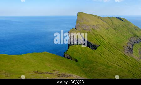 Torreggianti scogliere verdi si innalzano drammaticamente sopra il mare calmo, mostrando la bellezza naturale mozzafiato delle Isole Faroe. Una destinazione perfetta per amanti della natura e avventurieri. Foto Stock