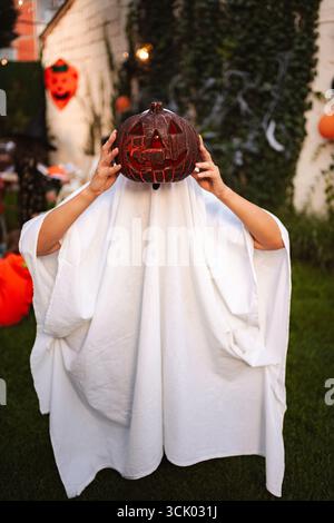 Bambino che indossa un costume fantasma che tiene in mano Jack-o'-lanterna durante la festa di Halloween nel cortile Foto Stock