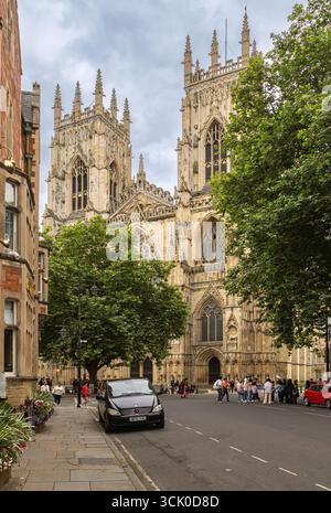Vista sulla strada della cattedrale di York in Inghilterra, con torri gotiche, vetrate colorate e sculture in pietra che si innalzano sopra i turisti su una strada alberata. Foto Stock