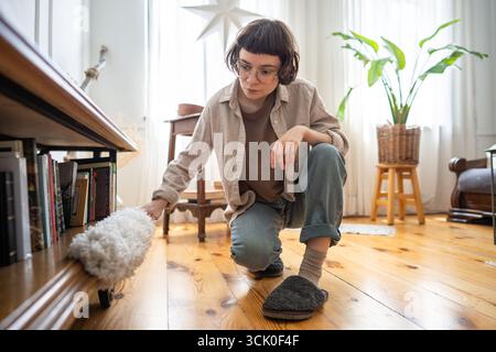 Una giovane donna ordinata che usa il filtro antipolvere per rimuovere la polvere dai mobili vicino al pavimento, routine di pulizia della casa Foto Stock
