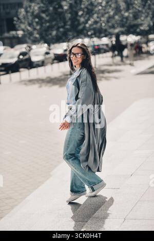 Donna sicura di sé con lunghi capelli marroni che cammina elegantemente su una strada cittadina con un abbigliamento casual che sfoggia fascino urbano e ritratti verticali Foto Stock