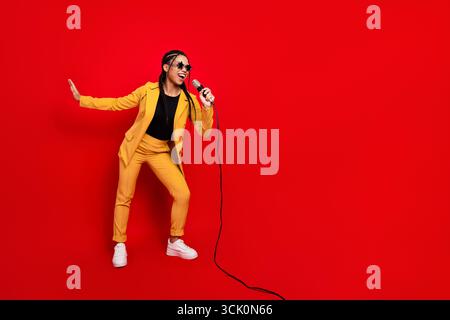 Giovane donna vibrante in tuta gialla che tiene il microfono e canta energicamente su uno sfondo rosso brillante Foto Stock