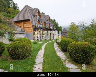 Fila di piccoli cottage in legno con tetti a punta, circondati da cespugli rifilati e sentieri in pietra. Turismo e alloggio in montagna in Serbia. Foto Stock