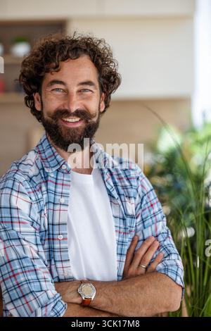 Ritratto della vista frontale di un uomo sorridente dai capelli ricci con baffi in piedi nel suo appartamento con le braccia incrociate Foto Stock