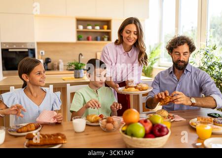 La famiglia si riunisce intorno a un tavolo in una cucina soleggiata, condividendo vari cibi per la colazione e godendosi del tempo di qualità insieme Foto Stock