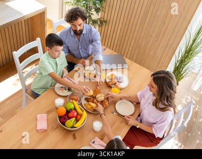 I membri della famiglia condividono un'allegra colazione con frutta, pasticcini e bevande seduti intorno al tavolo da pranzo in legno nella loro accogliente casa Foto Stock