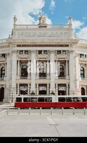 Vista dello storico Burgtheater di Vienna in una giornata di sole con un tram rosso che passa in primo piano, cielo blu con nuvole. Foto verticale. Foto Stock