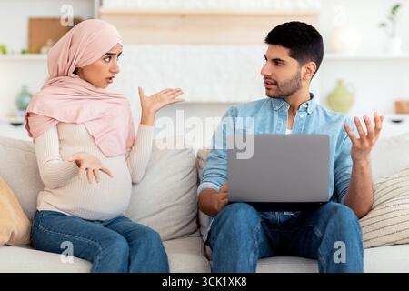 Stressata donna musulmana incinta gridando al marito mentre l'uomo usa il computer portatile, seduto insieme sul divano a casa Foto Stock