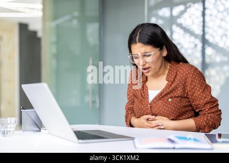 Giovane donna d'affari che soffre di gravi dolori addominali mentre è seduta alla scrivania in un ufficio moderno, aggrappandosi allo stomaco e soffrendo di disagio mentre lavora sul suo portatile Foto Stock