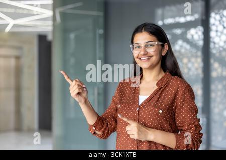 Donna d'affari sorridente che indossa gli occhiali, punta con le dita indice in uno spazio vuoto in un ufficio luminoso e moderno, invita a comunicare e mostra professionalità Foto Stock