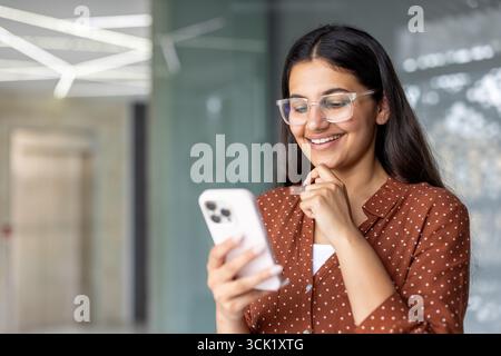 Donna d'affari felice che indossa gli occhiali da vista e utilizza il suo telefono cellulare, sorride brillantemente mentre si trova in un corridoio dell'ufficio, godendosi una meritata pausa durante la sua intensa giornata di lavoro Foto Stock