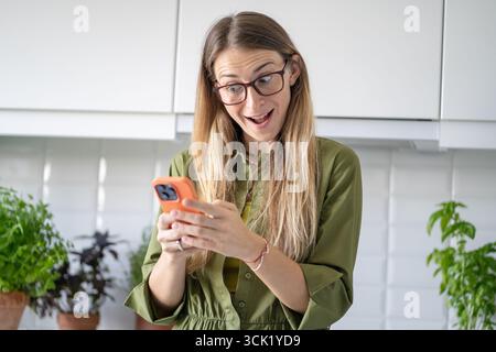 Donna stupita con smartphone che riceve buone notizie. Chiacchierate e videochiamate femminili a casa Foto Stock