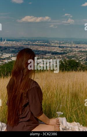La ragazza con la camicia color cioccolato e i lunghi capelli marroni si siede su una coperta in mezzo all'erba verde-gialla alta, tenendo in mano un bicchiere di vino bianco. Vista panoramica di Foto Stock
