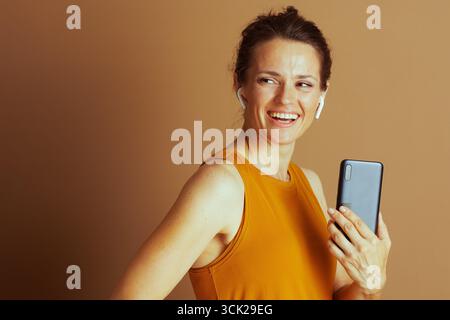 Una donna tra gli anni '30 e '40 con un panino, che indossa la parte superiore gialla della senape, sorride in grande mentre ascolta l'audio attraverso gli auricolari e tiene in mano uno smartphone, truffa Foto Stock