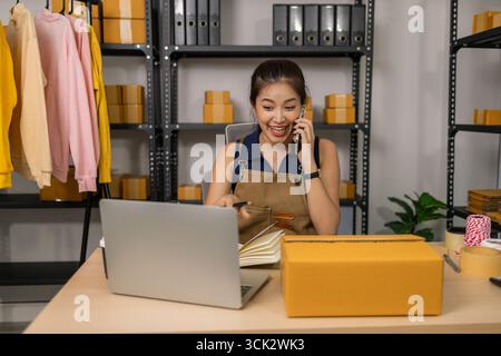 Giovane imprenditrice sorridente che parla al telefono cellulare e lavora al computer portatile dall'ufficio domestico, gestendo gli ordini dei clienti per il suo abbigliamento online Foto Stock