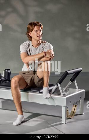 Un giovane seduto su un riformatore Pilates, vestito con abbigliamento casual, in uno studio ben illuminato. Sembra rilassato e contemplativo, incarnando bene Foto Stock