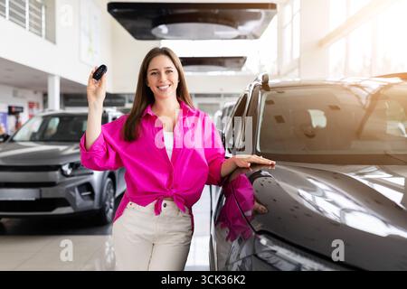 Giovane donna felice che posa con le chiavi dell'auto accanto al veicolo nuovo presso la concessionaria Foto Stock