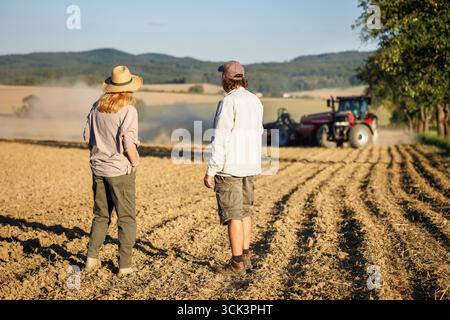 Lavoro di squadra nelle zone rurali con gli agricoltori che guardano al trattore e alla seminatrice che preparano il terreno per la coltivazione. Agronomo che controlla l'attività di semina in campo agricolo Foto Stock