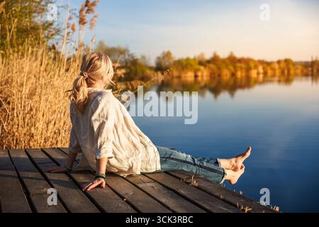 Donna seduta e rilassante a piedi nudi sul molo di legno vicino al lago durante l'ora d'oro dell'estate indiana Foto Stock