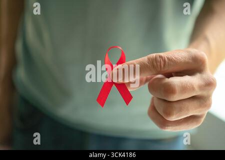Primo piano di una mano maschile che regge il nastro rosso di raso, simboleggiando l'HIV AIDS, supporta il ricordo e la speranza per una cura futura durante il mese di sensibilizzazione di dicembre. Foto Stock