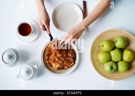 Top down di donna che taglia la torta con il coltello al tavolo, mani visibili vicino al piatto con mele verdi, tazza di tè e teiere disposte in superficie, vista dall'alto composizione Foto Stock