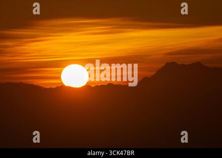 Vista del sole che si illumina intensamente mentre si innalza sulla sagoma scura di montagne lontane sullo sfondo di nuvole arancioni ardenti, Sajek, Chittagong Division, Bangladesh. Foto Stock